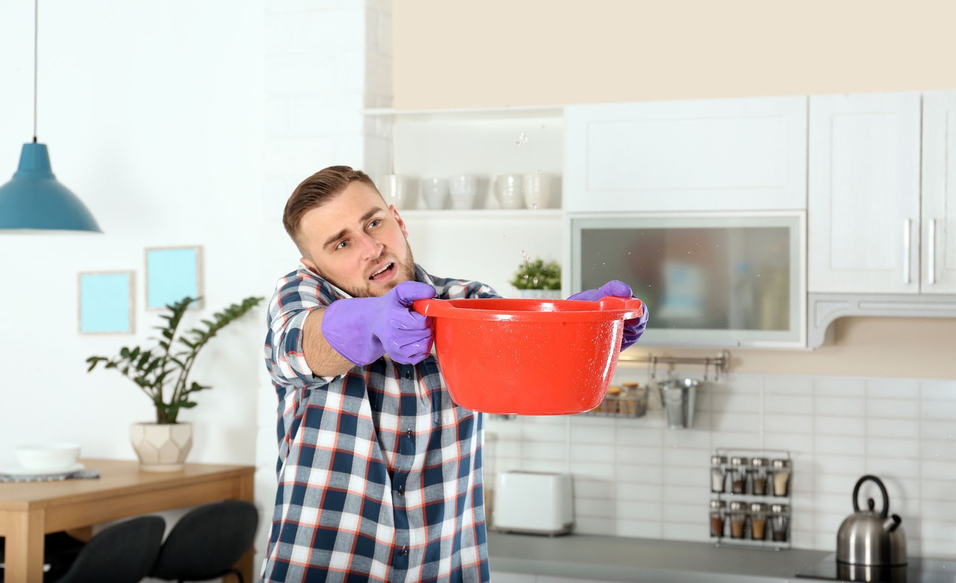 Man in plaid shirt and purple gloves, holding a red bowl to catch water leaking from a ceiling in a kitchen.