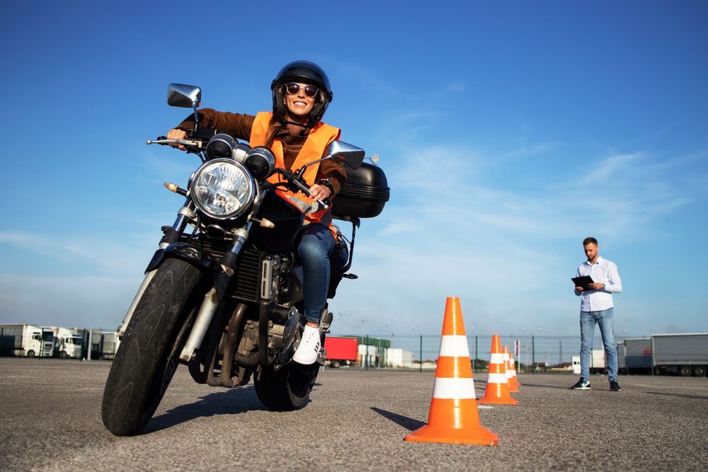 Woman on motorcycle taking a driving test. She is smiling, wearing a helmet and orange vest; an instructor watches.