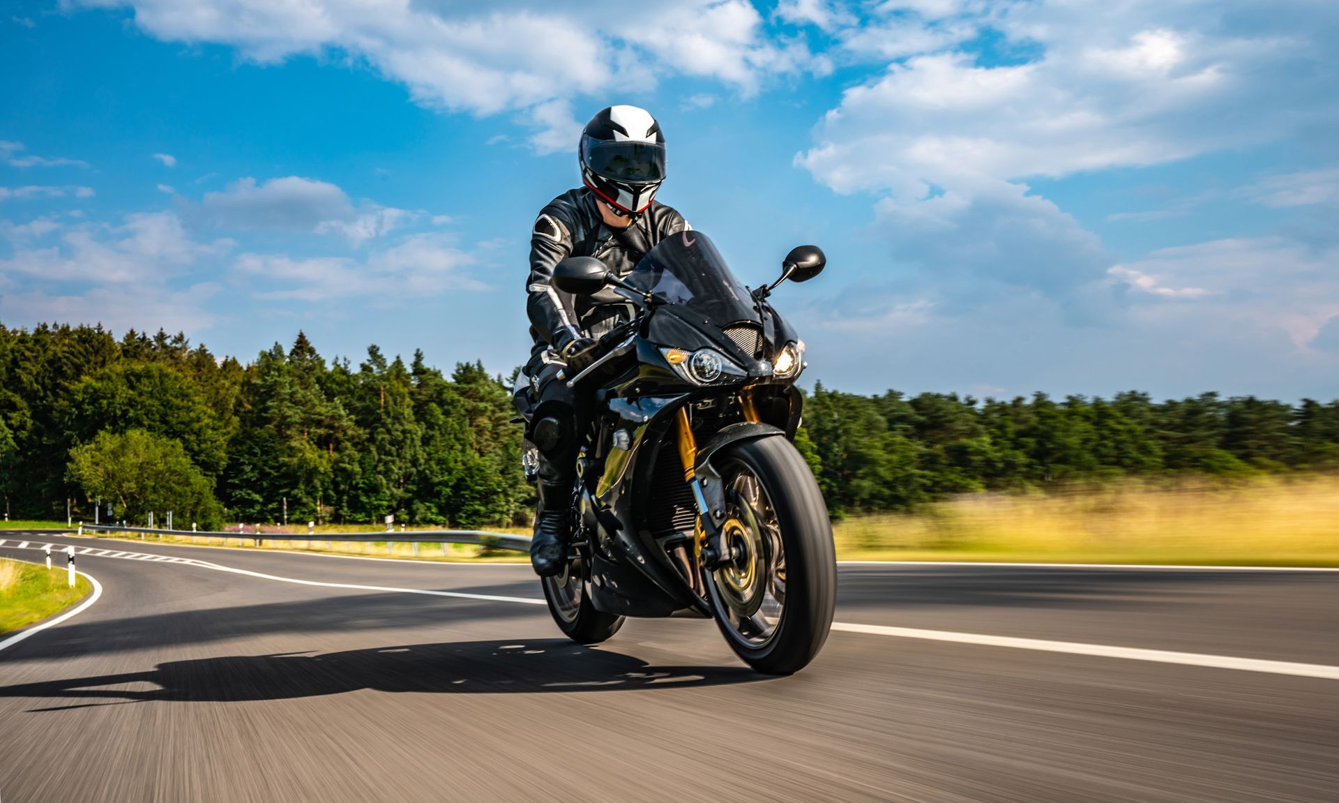 Motorcyclist in black leather riding on a curved road, sunny day.
