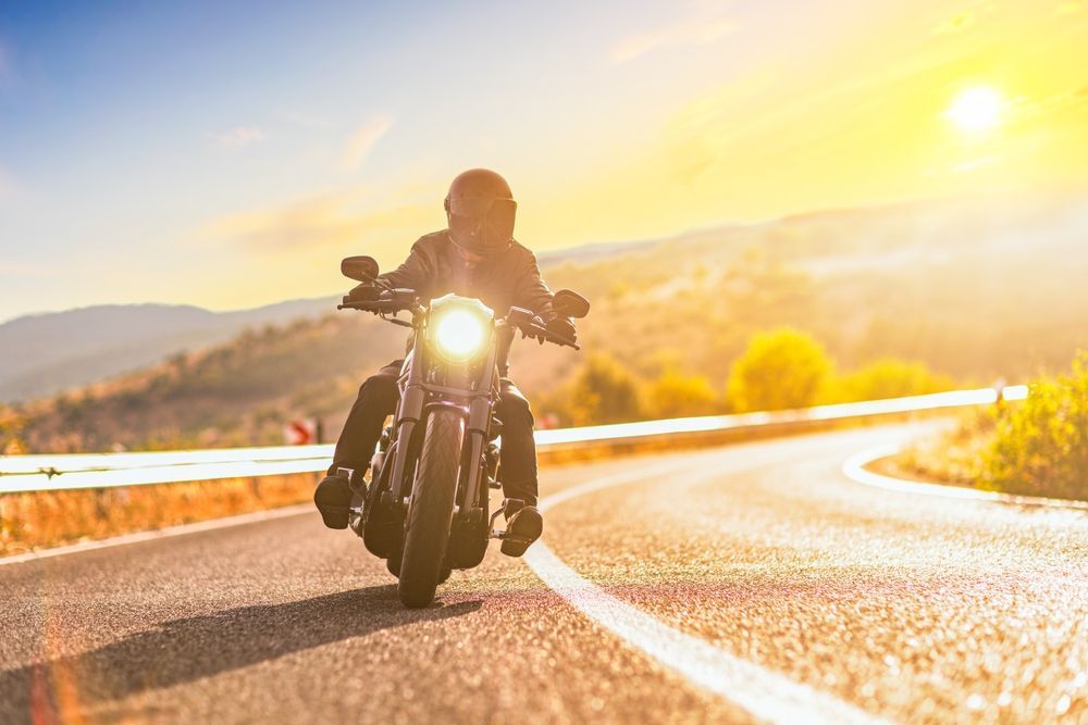 Motorcyclist riding on a curved road, bright sun, mountains in the background.