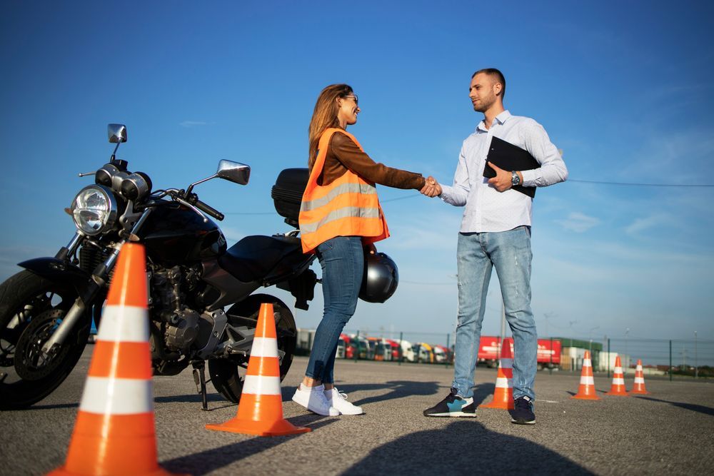 Woman and man shake hands next to a motorcycle and cones outdoors on a sunny day.