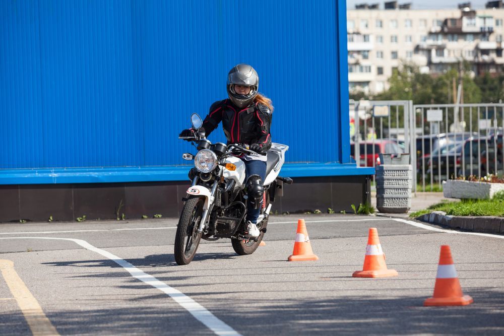 Person on motorcycle navigating an obstacle course marked by orange cones in an open, paved area.