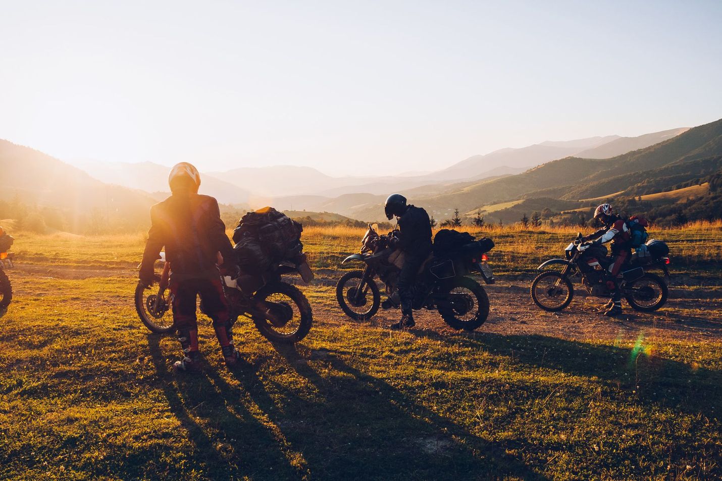 Motorcyclists on a dirt path in mountains at sunset.  One rider stands by bike; others start.  Golden light.