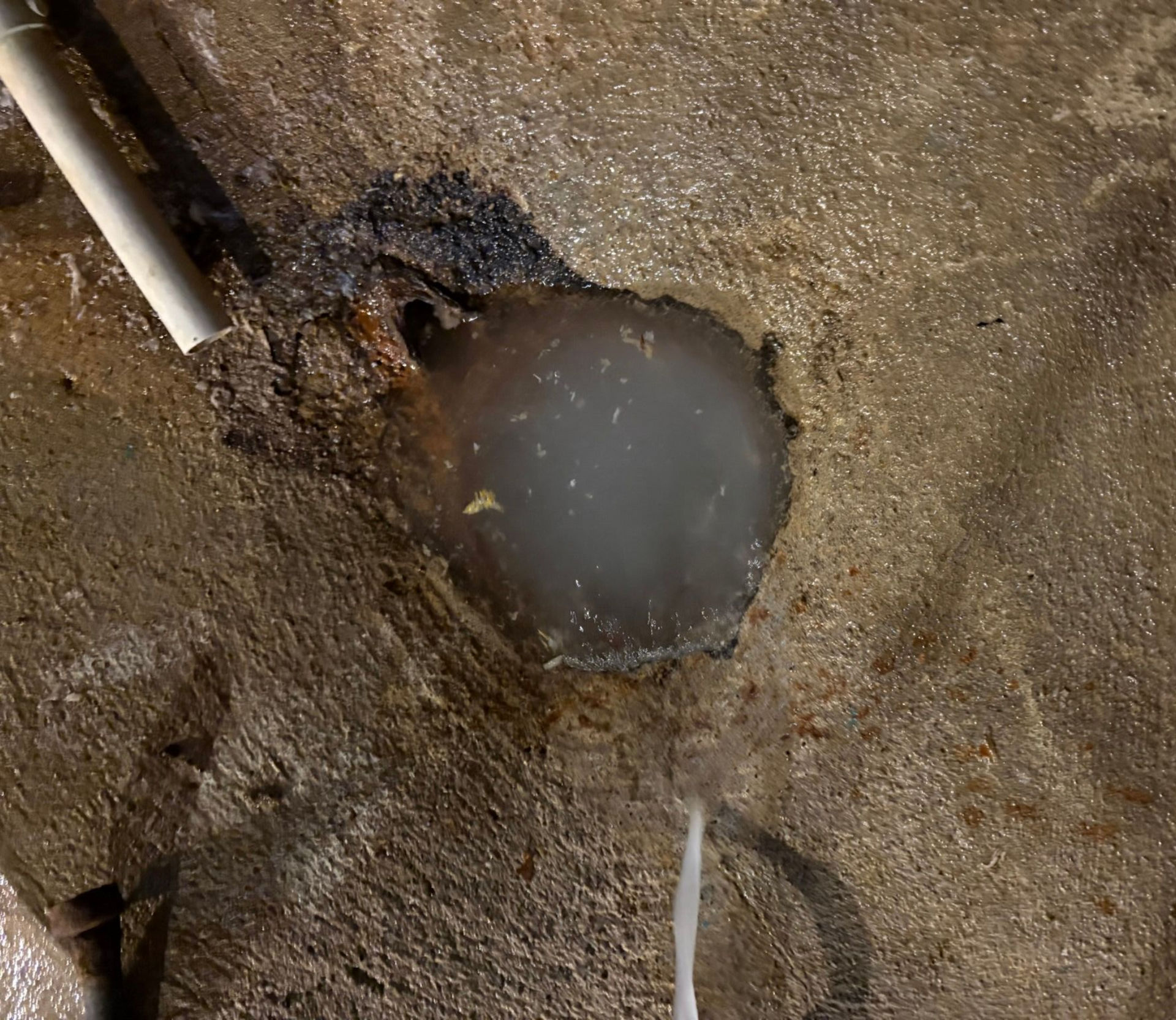 Round hole in dirty concrete with standing water and a pipe at the upper left
