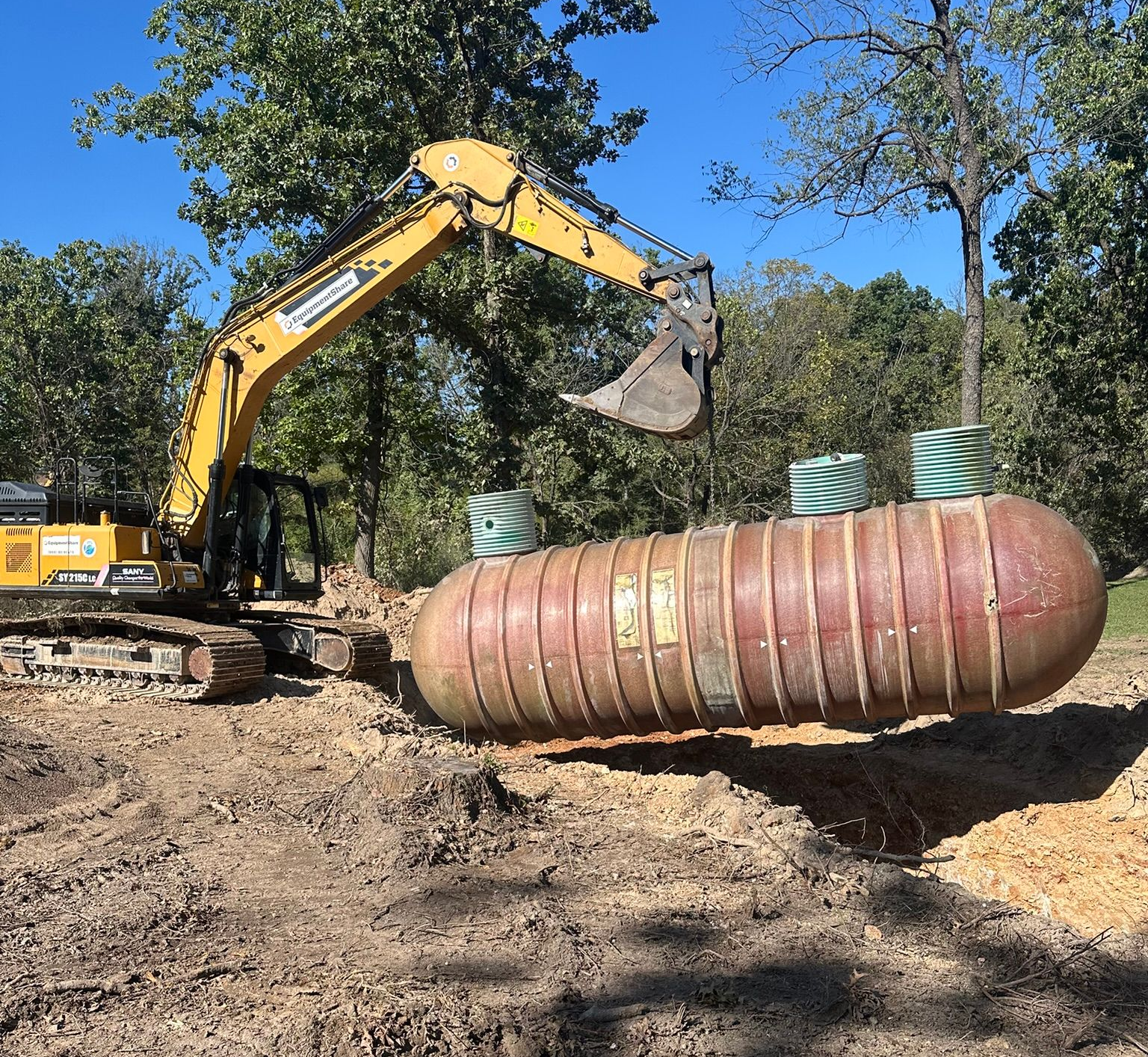Excavator lifting a large rust-colored metal tank at a dirt construction site, with trees in the background
