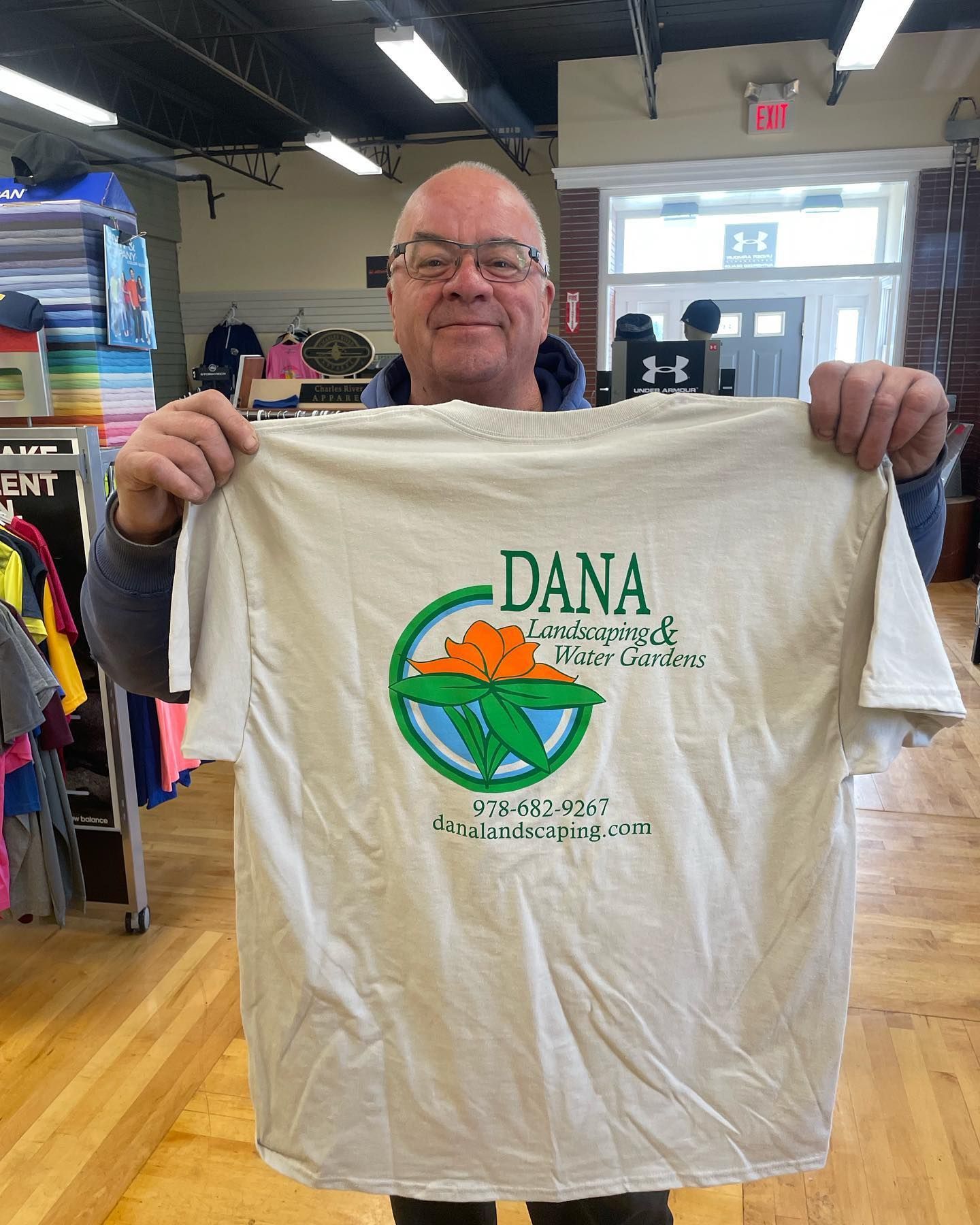 Man holding a tan t-shirt with a green and orange logo; inside a shop with clothing racks.