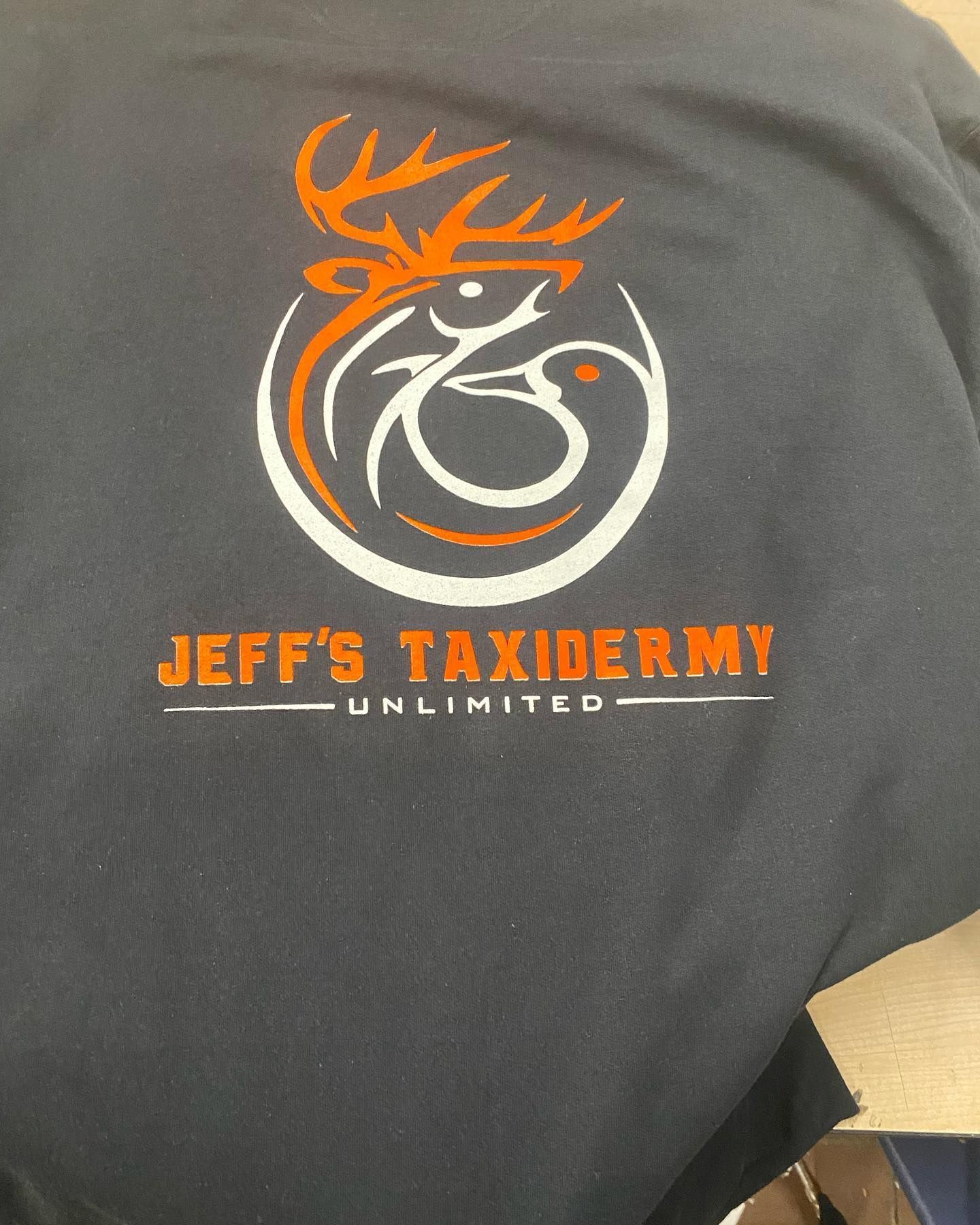 Logo on a dark t-shirt for Jeff's Taxidermy. The logo features a deer head, duck, and text in orange and white.