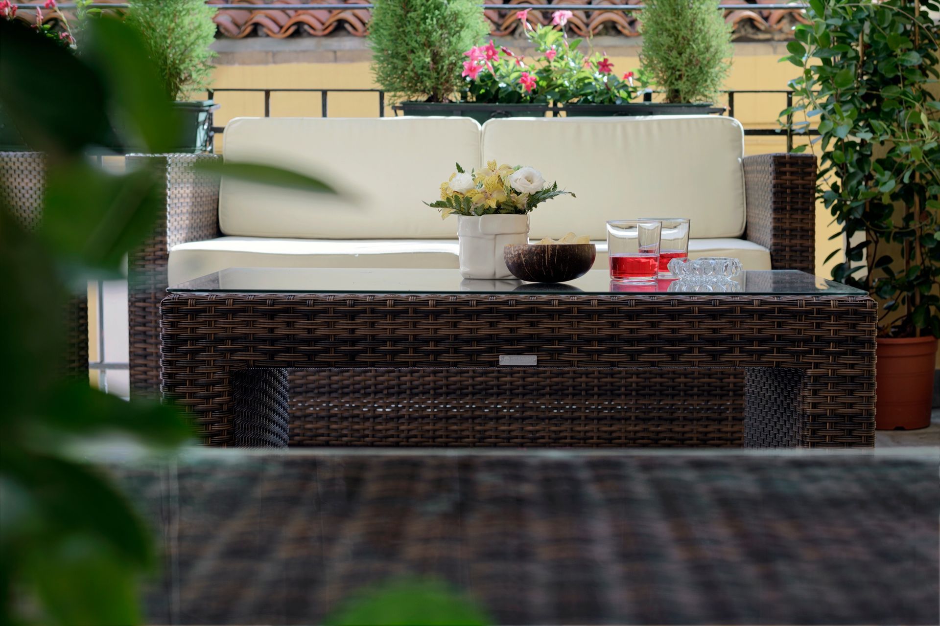 Terrasse extérieure avec canapé en osier, table basse, coussins et plantes en pot contre un mur couleur crème.