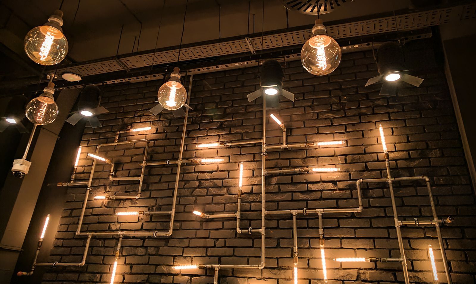 Brick wall with copper pipes forming a lit pattern, illuminated by glowing bulbs and spotlights.