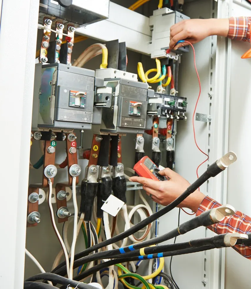 Hands wiring electrical panel with a green tool, numerous wires and circuit breakers visible.