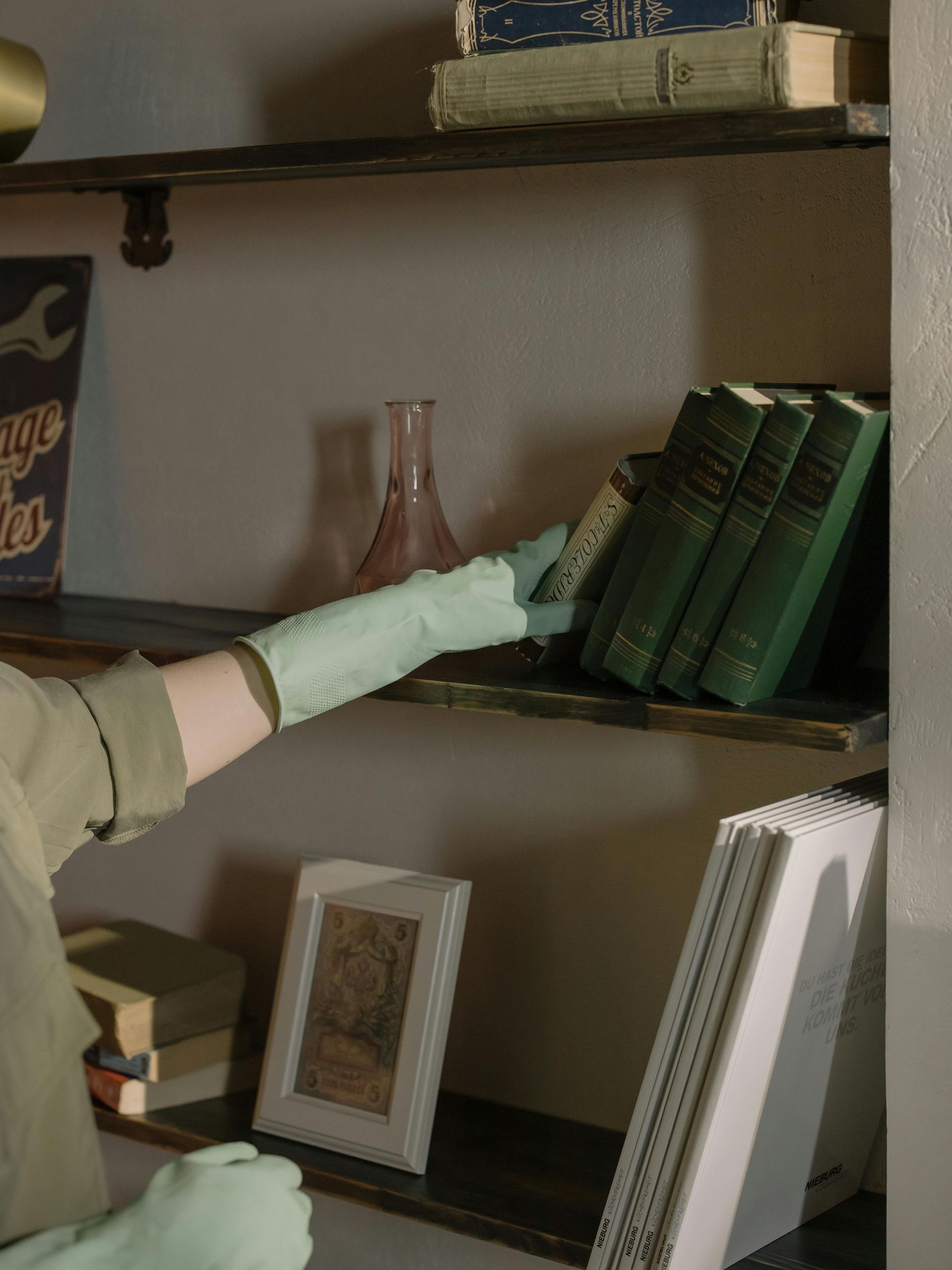 Gloved hand reaching for green books on a shelf beside a pink vase and framed photo