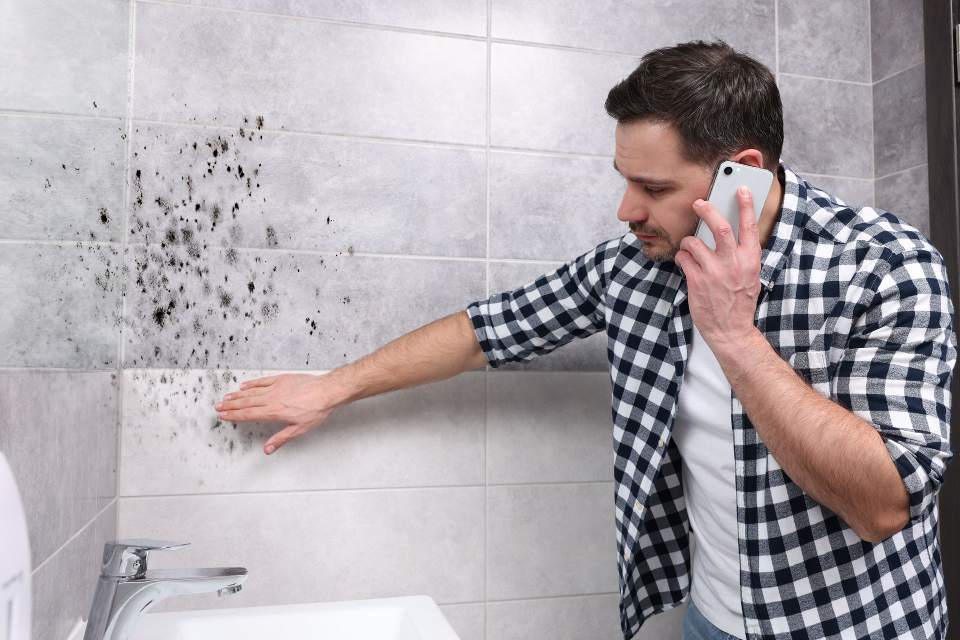 Person inspecting black mold on bathroom tiles while making a phone call near a sink.