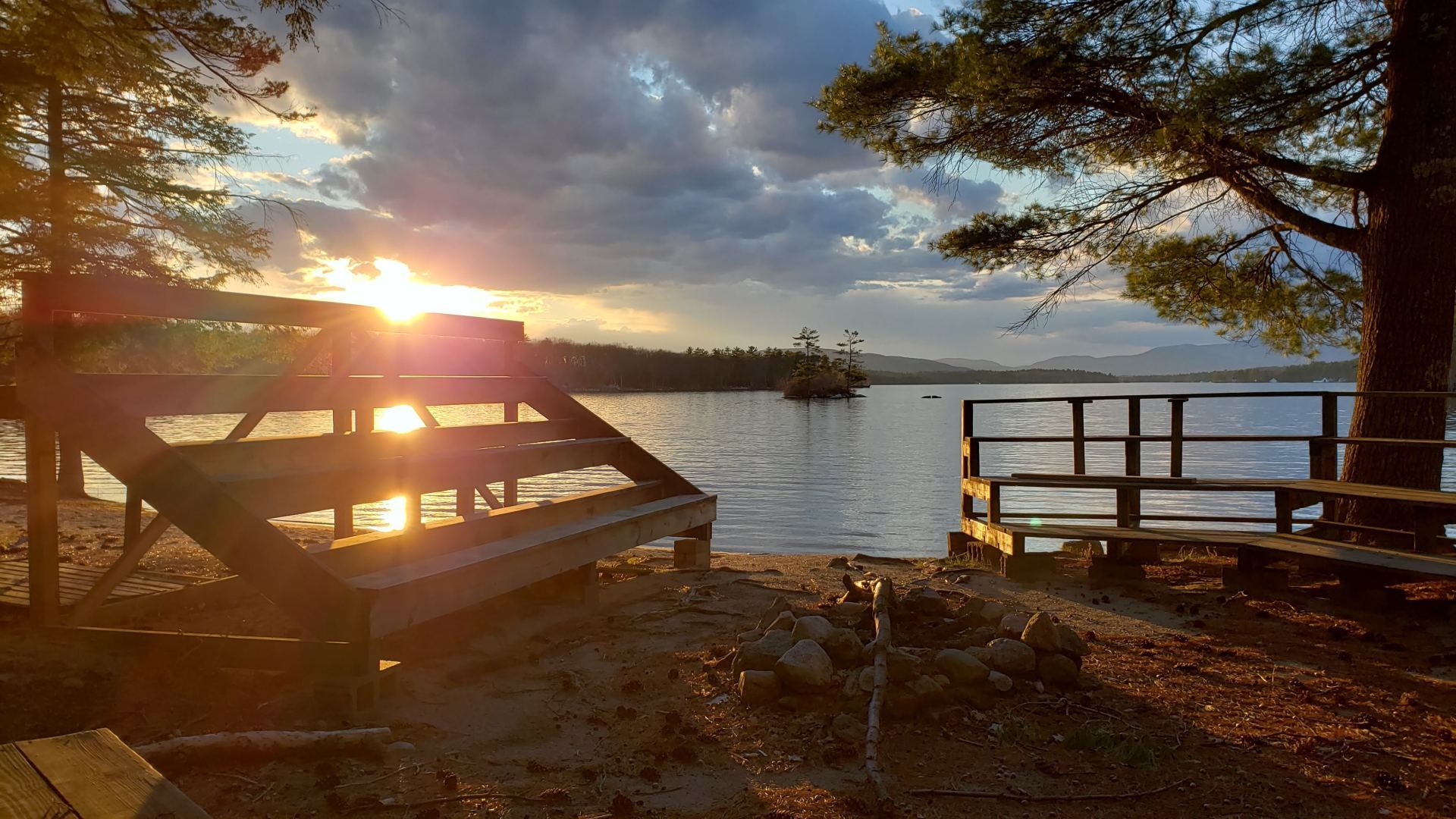 The sun is setting over a lake with a picnic table in the foreground.