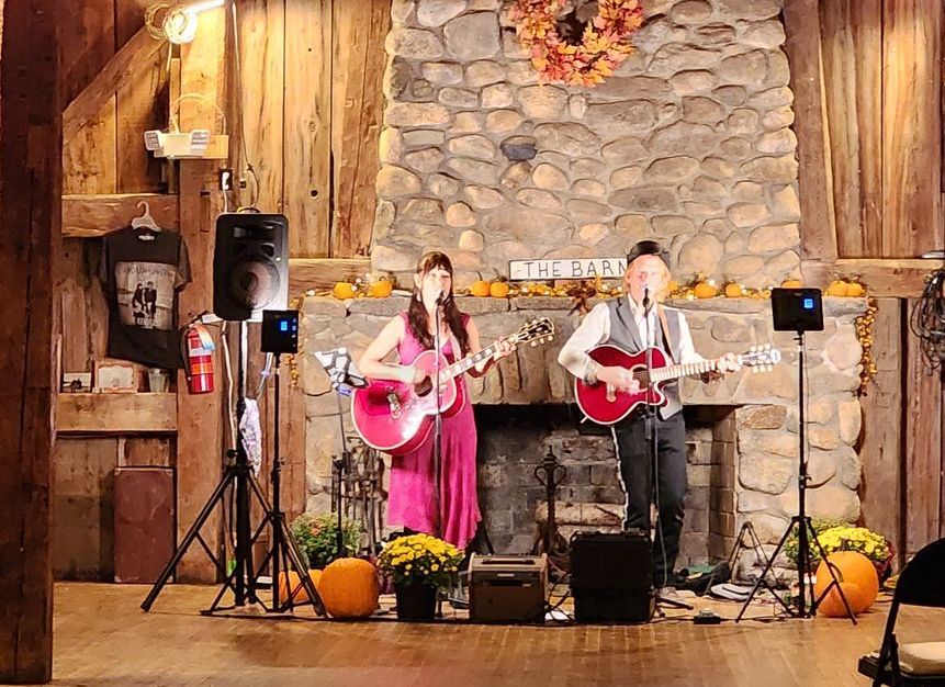 A man and a woman are playing guitars and singing in front of a fireplace.