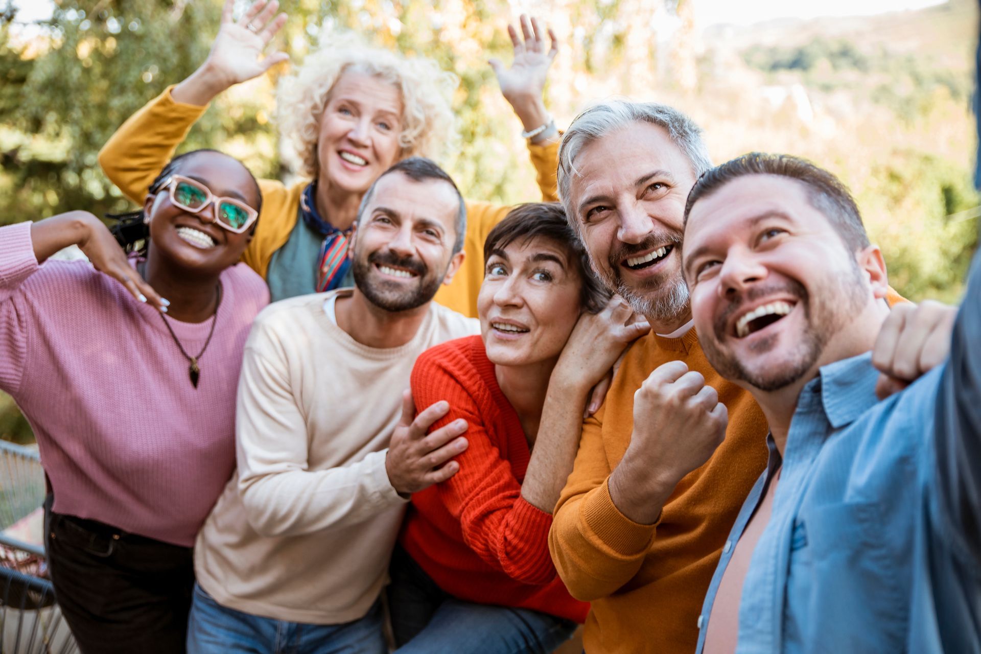Group of smiling adults taking a selfie outdoors; diverse ethnicities, happy expressions, waving.