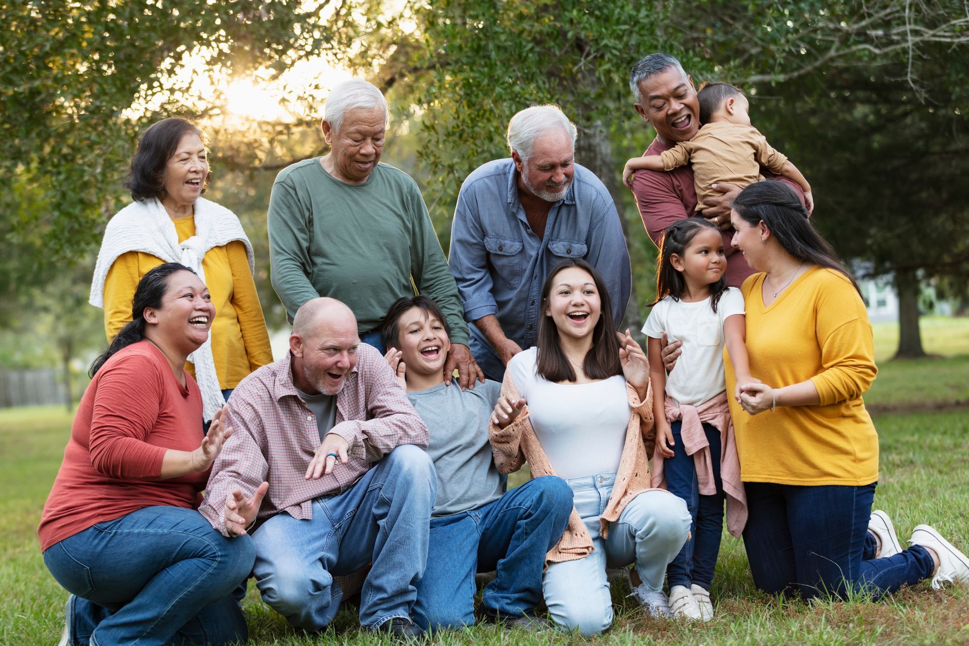 Large multigenerational family smiles, posing outdoors. People of various ages laugh and embrace in a grassy area.