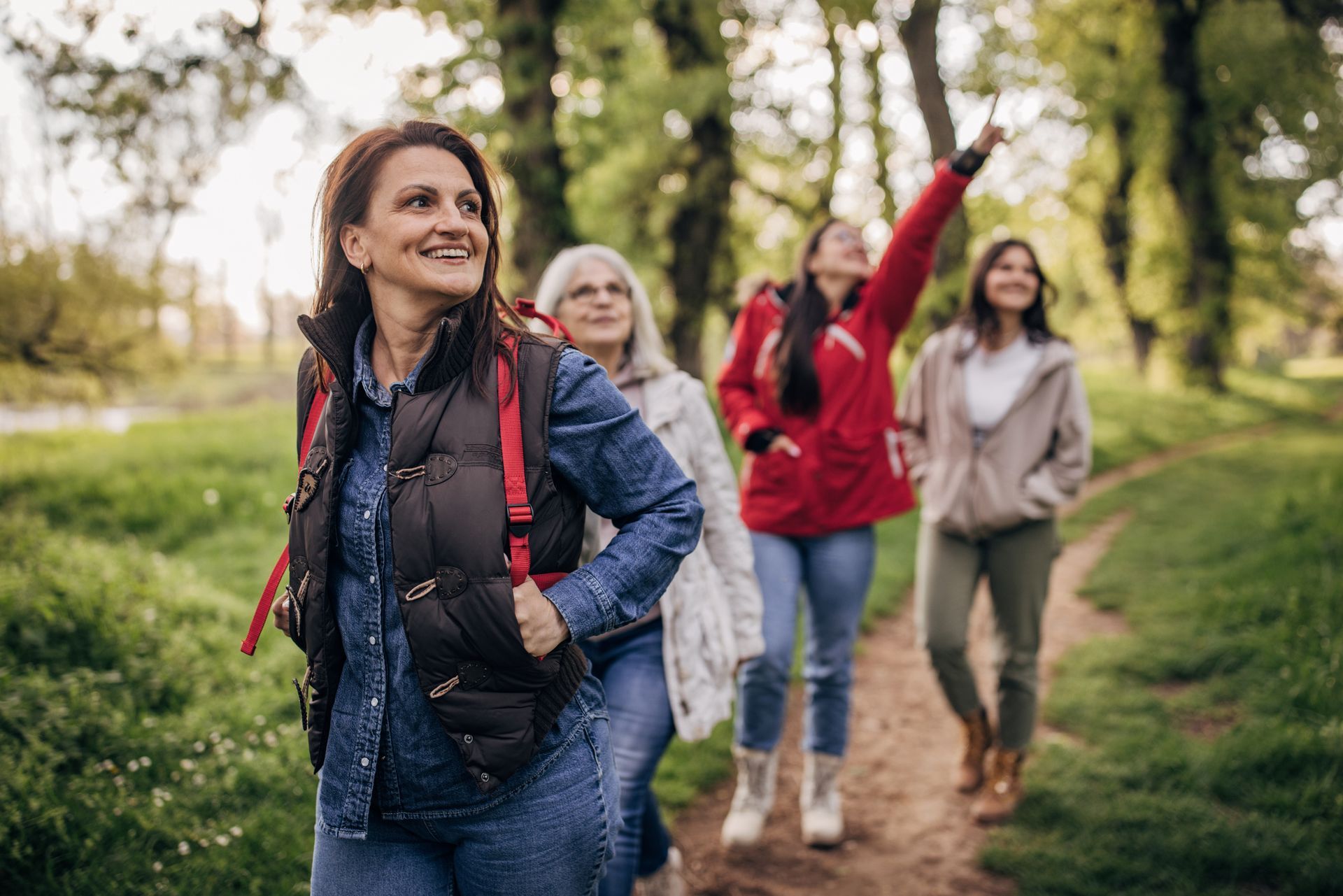 Four women hike on a path in a park, one points up, all smiling.