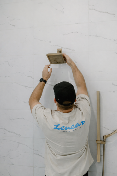 A person wearing a t-shirt and black cap attaches a rectangular shower head to a white marble-tiled wall.