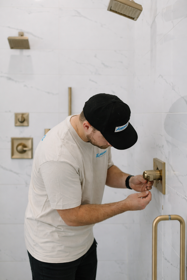 A person wearing a black cap and beige t-shirt installing gold shower hardware on a white marble tiled wall.
