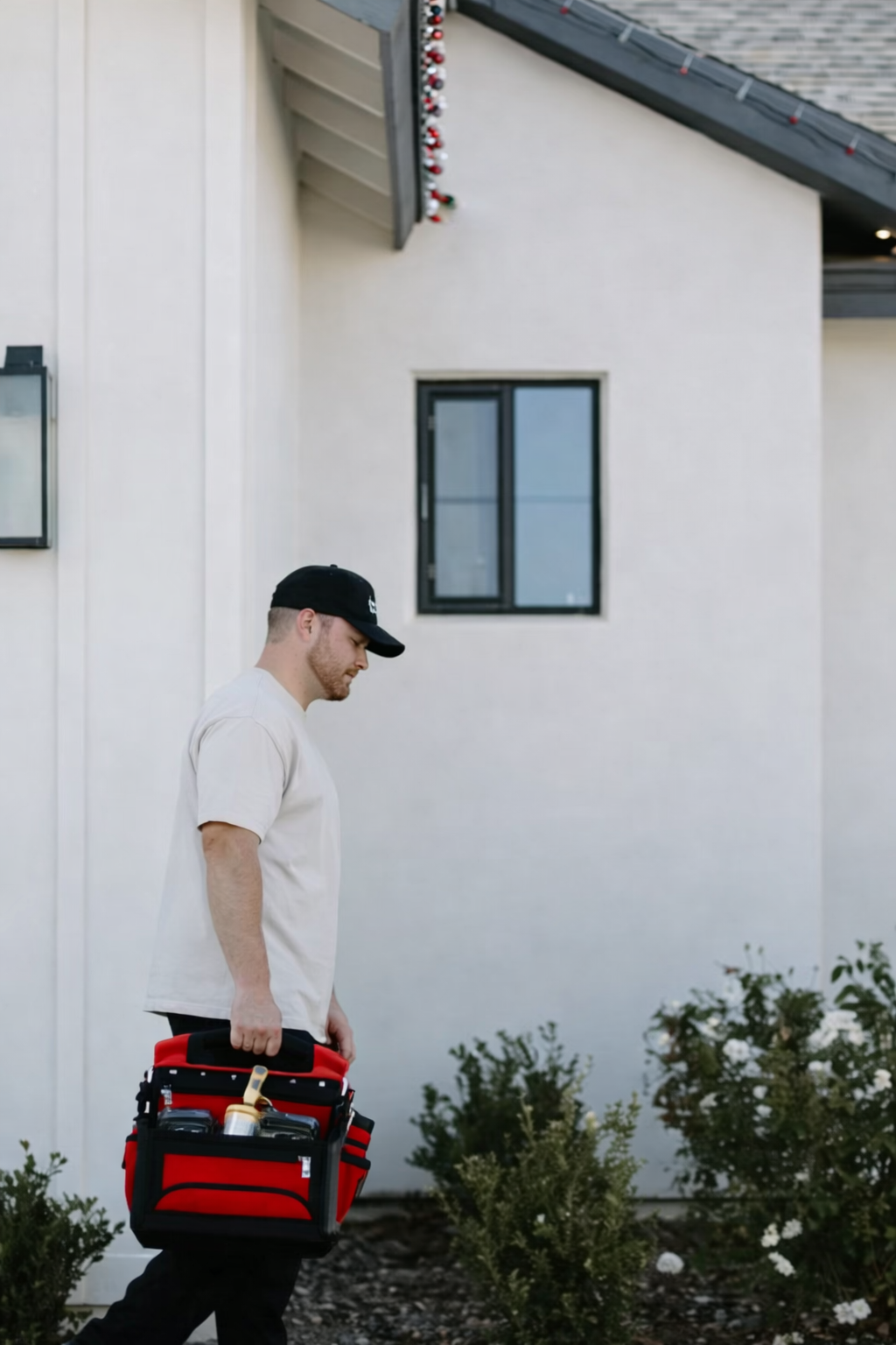 A person in a neutral t-shirt and cap walks toward a white house while carrying a red tool bag.