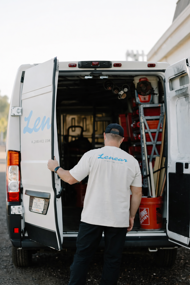 A person in a logo t-shirt stands at the open rear doors of a white work van filled with tools and equipment.