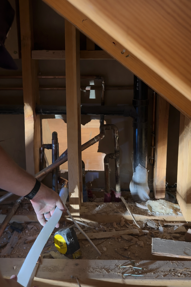 A person’s hand holds a white measuring tape in a dark, unfinished crawl space with exposed wooden studs and plumbing.