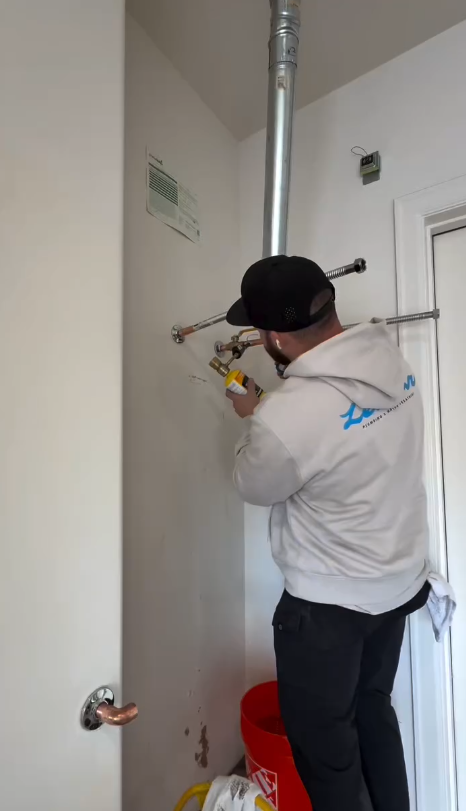 A person in a gray hoodie and black cap installs pipes into a white wall inside a room, holding a yellow tool.