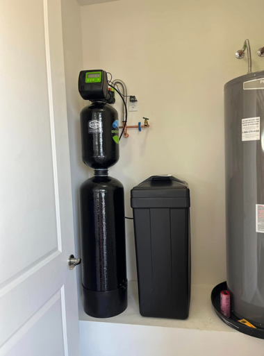 A black water softener system and tall brine tank placed next to a water heater in a closet.