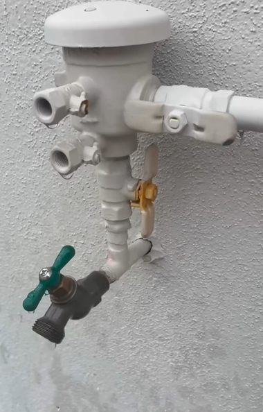 A wall-mounted backflow prevention device with a faucet and control valves, painted white, against a textured wall.