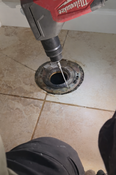 A person uses a red Milwaukee drill to bore a hole through a metal toilet flange set into a tiled bathroom floor.