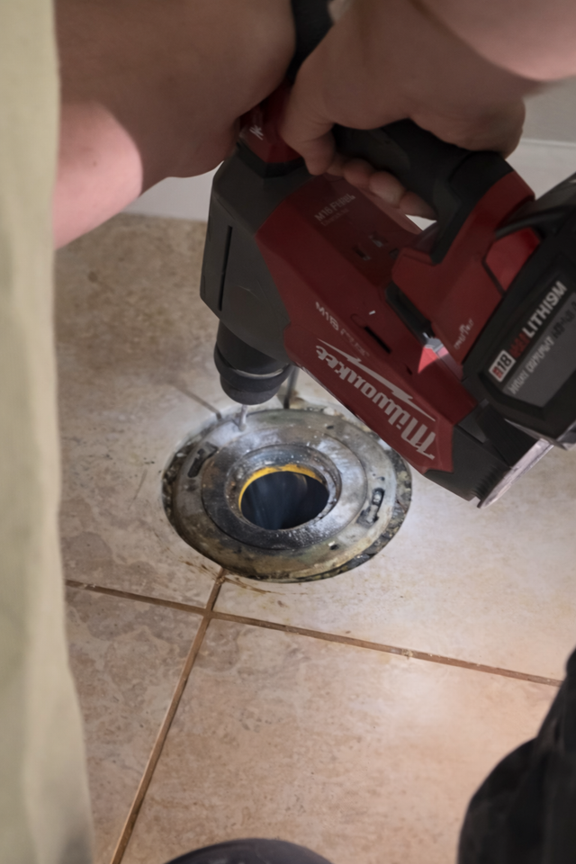 A person uses a red Milwaukee power drill to screw a metal toilet flange into a tiled bathroom floor.