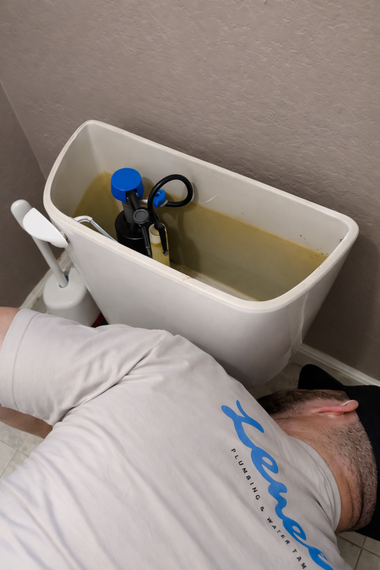 A plumber wearing a branded uniform works on the open, water-filled tank of a white ceramic toilet.