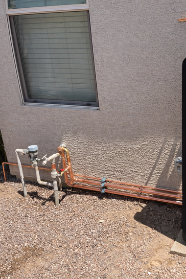 Residential exterior wall featuring an exposed copper plumbing manifold, a water meter, and pipe connections on gravel.