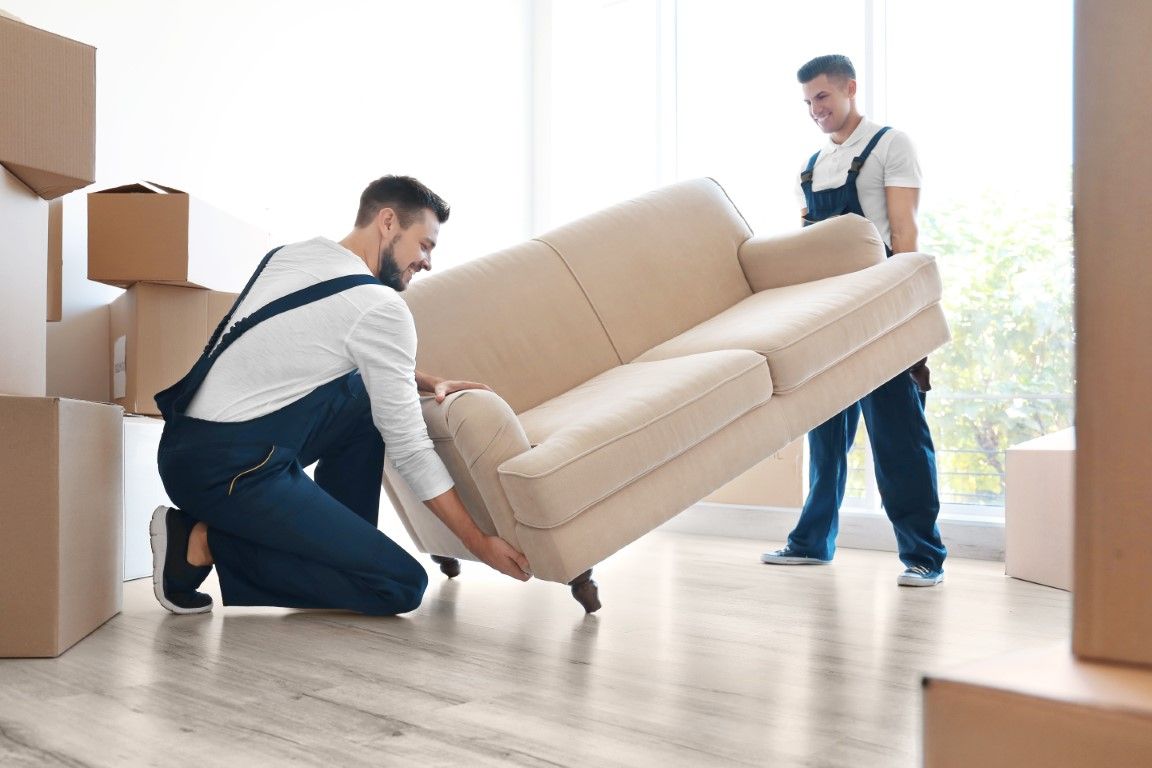 Two movers carrying a beige sofa through a bright room with stacked moving boxes