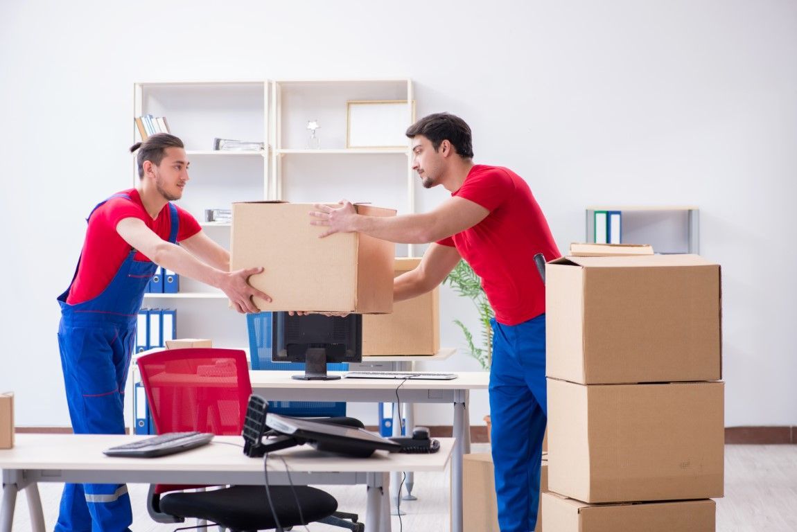 Two movers in red shirts carrying a cardboard box in a bright office beside stacked boxes