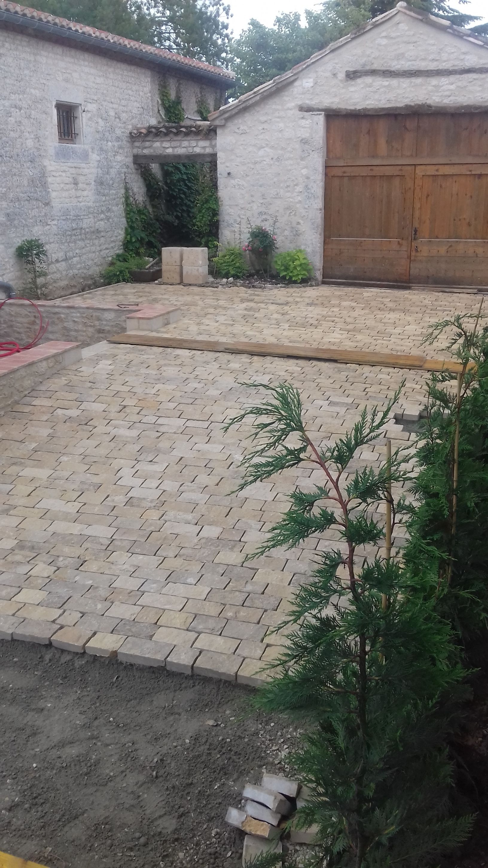Cour pavée jouxtant un bâtiment en pierre et une porte de garage en bois, avec une petite plante en pot.