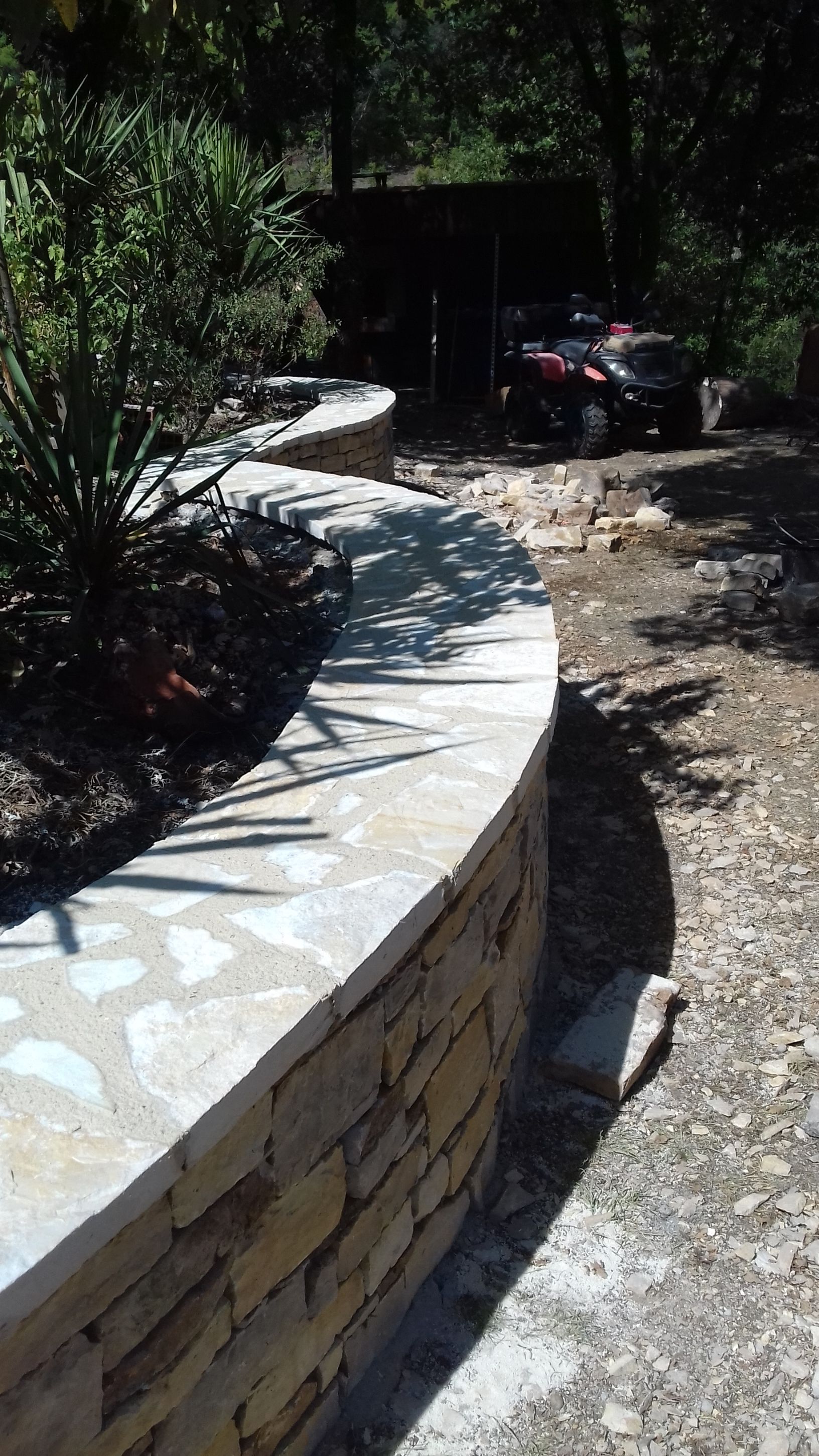 Banc en pierre incurvé le long d'un chemin de jardin ombragé, baigné de soleil et entouré d'une végétation dense.