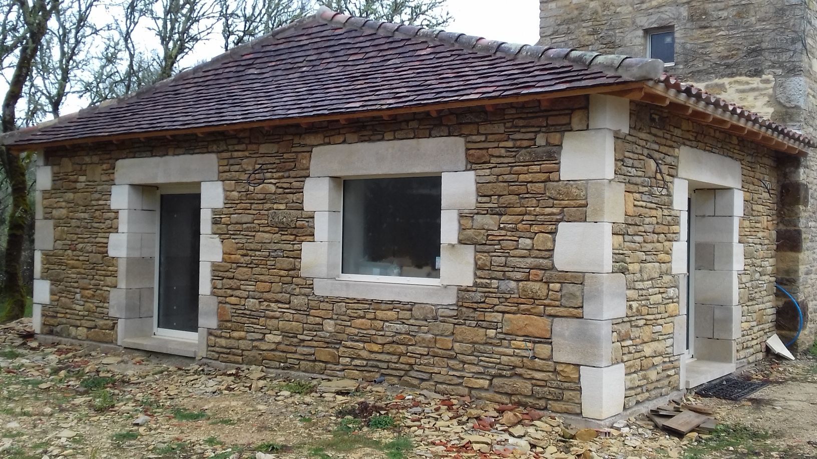 Petit cottage en pierre au toit de tuiles rouges et aux fenêtres à encadrement blanc, vu du coin extérieur