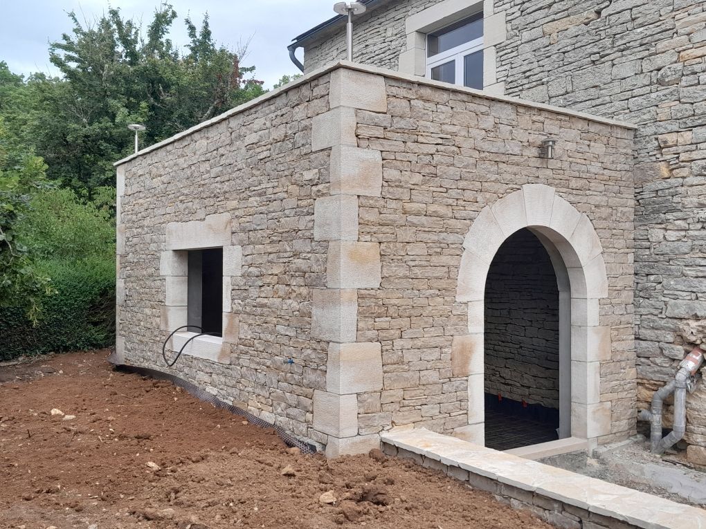 Maison en pierre avec porte cintrée et petite fenêtre, située à côté d'un bâtiment en pierre plus grand.