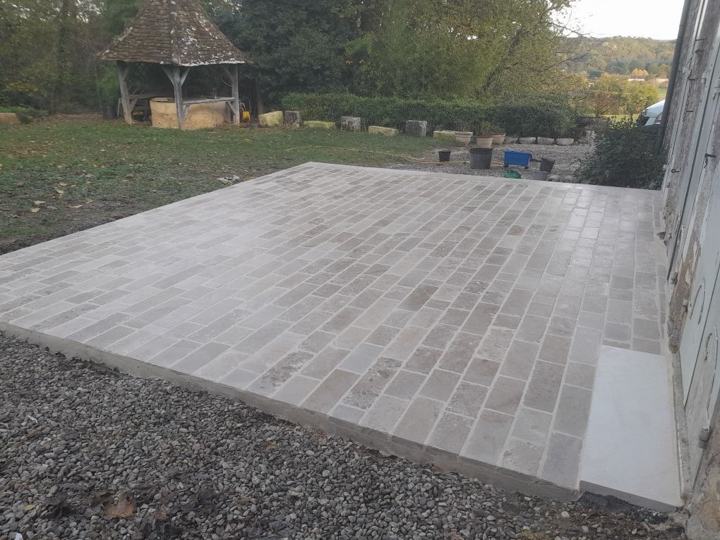 Terrasse en pierre jouxtant une maison, bordée de gravier et d'un jardin avec un petit kiosque en arrière-plan.