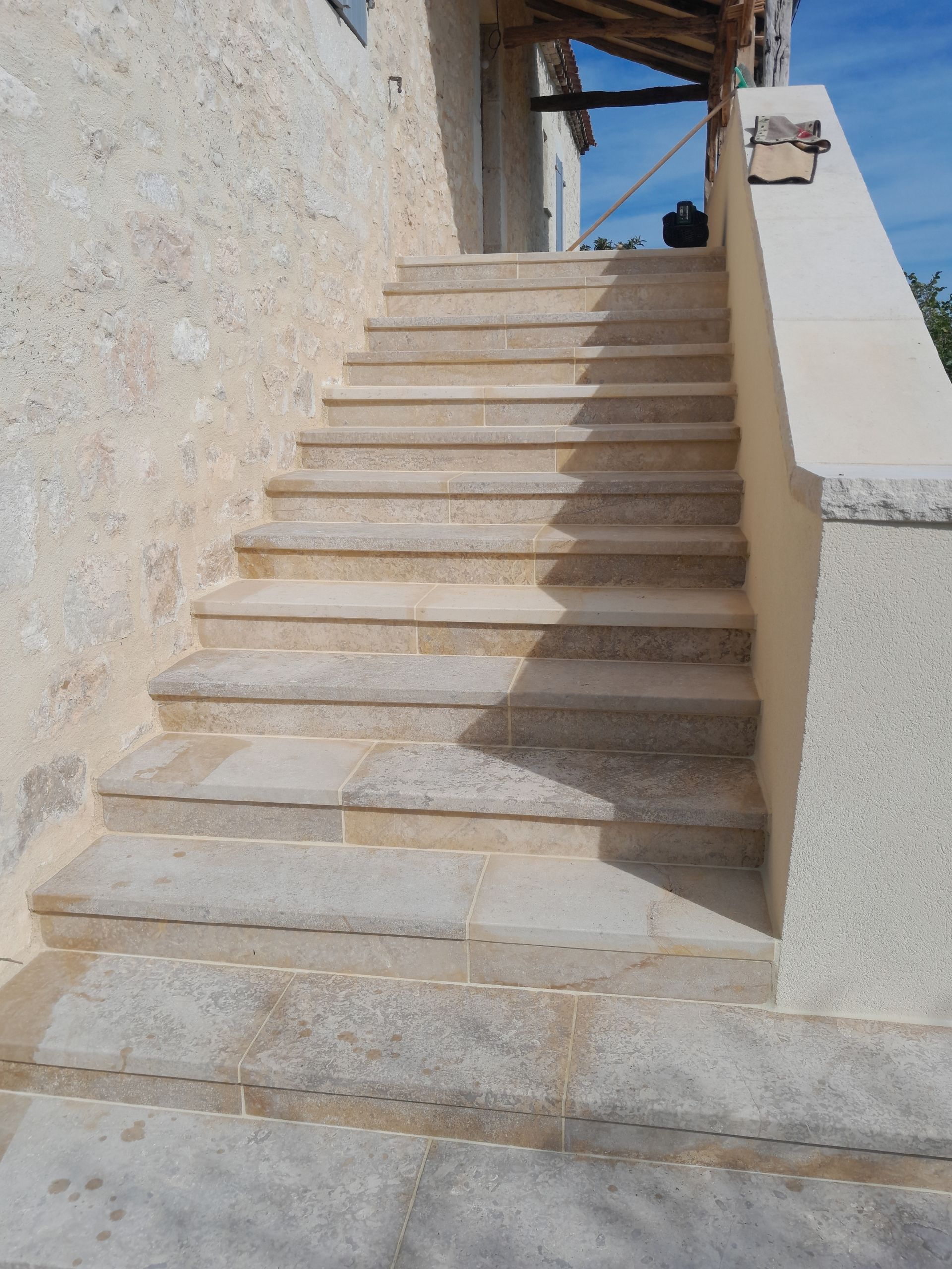 Un escalier en pierre longeant un mur beige et mène à une terrasse extérieure sous un ciel d'un bleu éclatant.