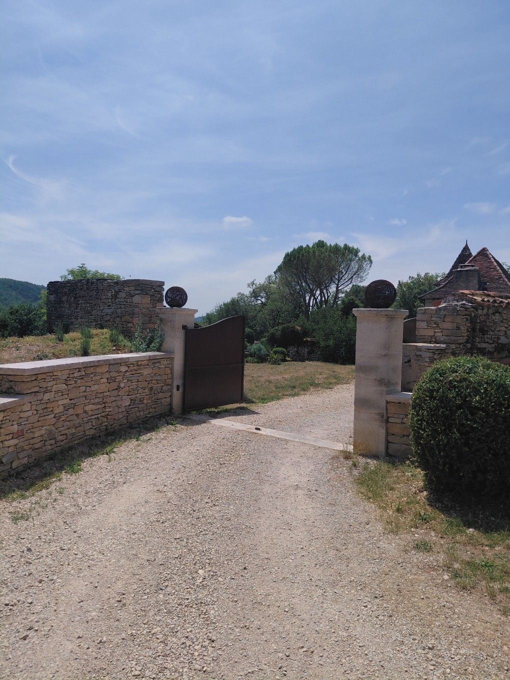 Un chemin de gravier mène, par un portail en pierre, à un jardin ensoleillé parsemé de ruines et d'arbres sous un ciel bleu.
