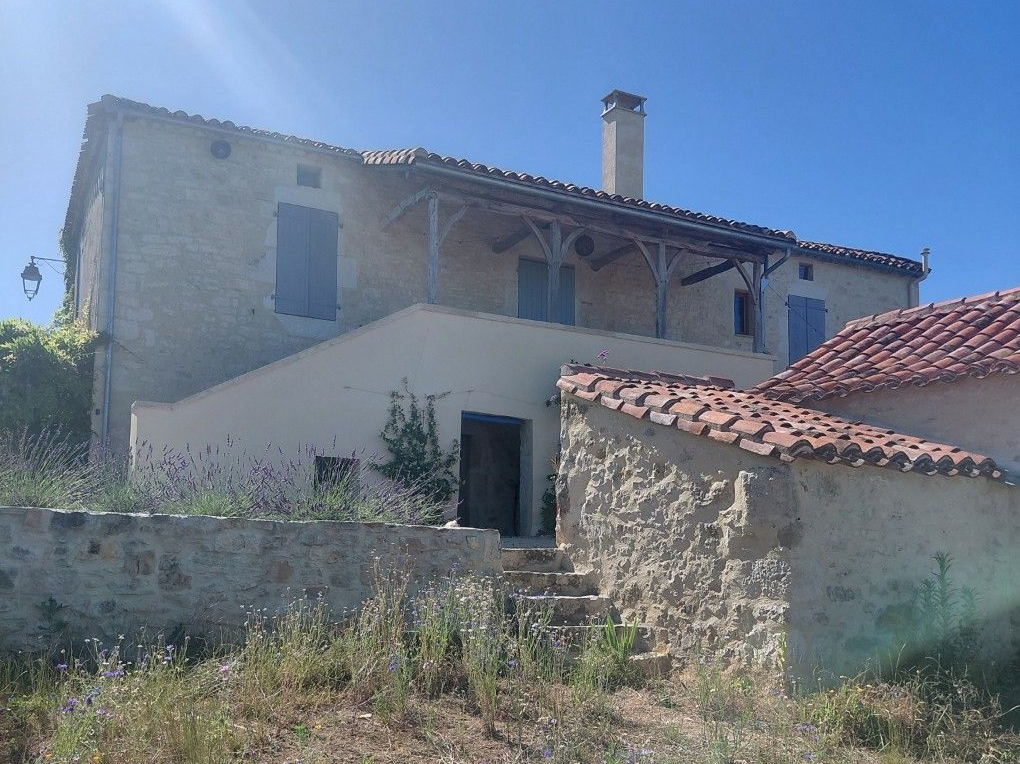 Ferme en pierre au toit de tuiles rouges et aux murs blancs, située derrière un muret en pierre dans une zone rurale ensoleillée