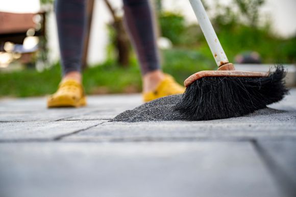 Person sweeping gray sand from between paving stones with a broom, wearing yellow shoes.
