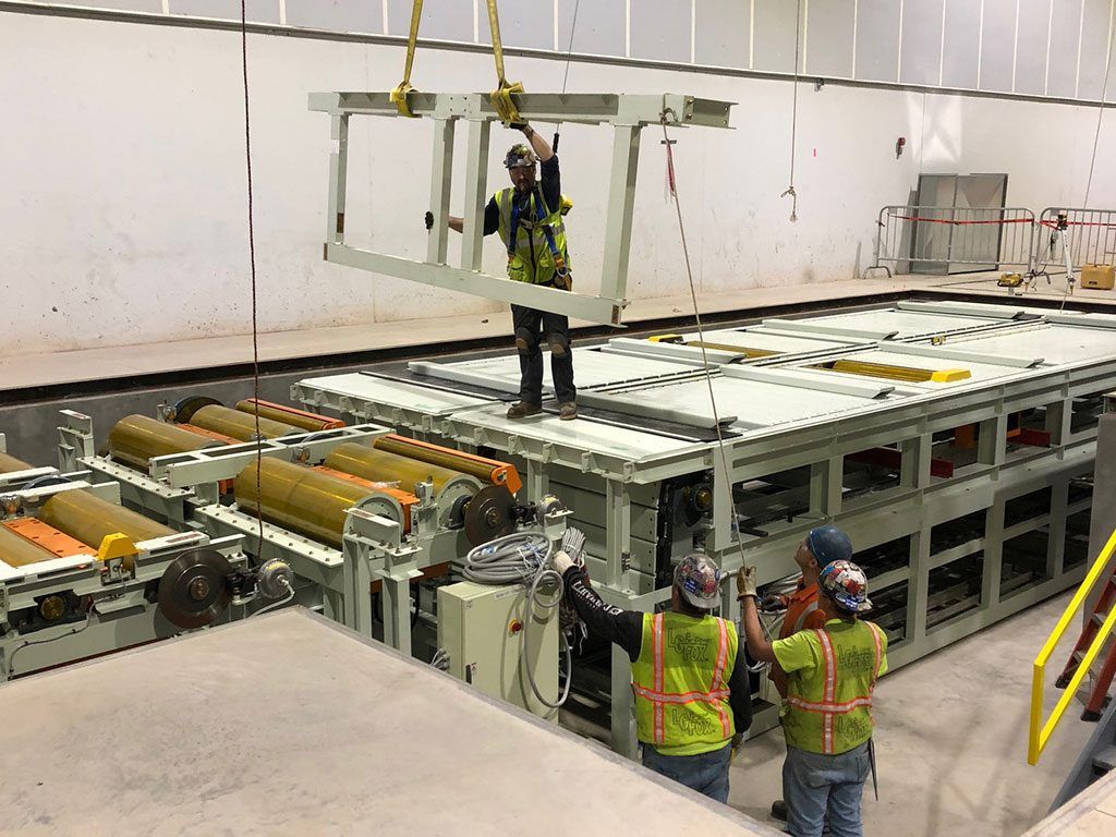Workers installing a rectangular metal frame suspended above machinery in a warehouse.