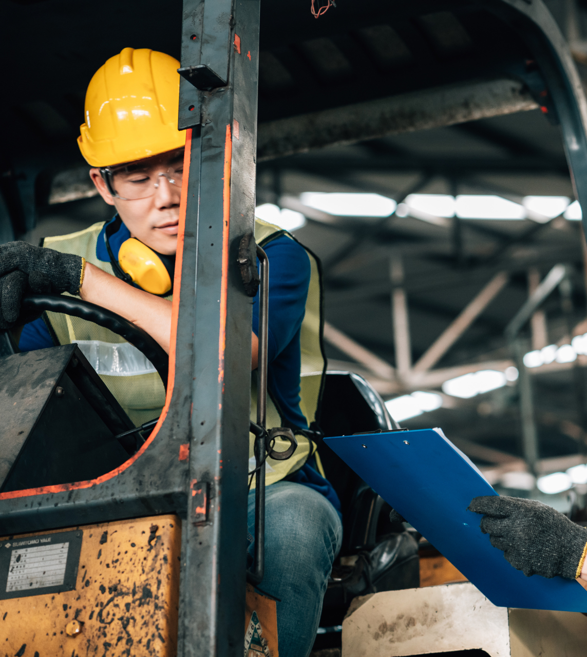 Person in hard hat operating a forklift, looking at a clipboard in a warehouse.