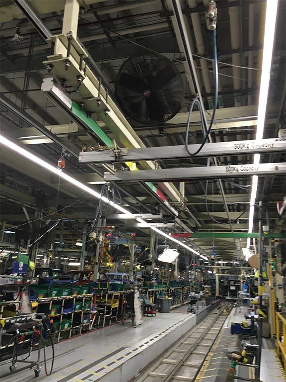 Assembly line in a factory. Overhead rails, lights, and a large fan. Parts and equipment along the line.