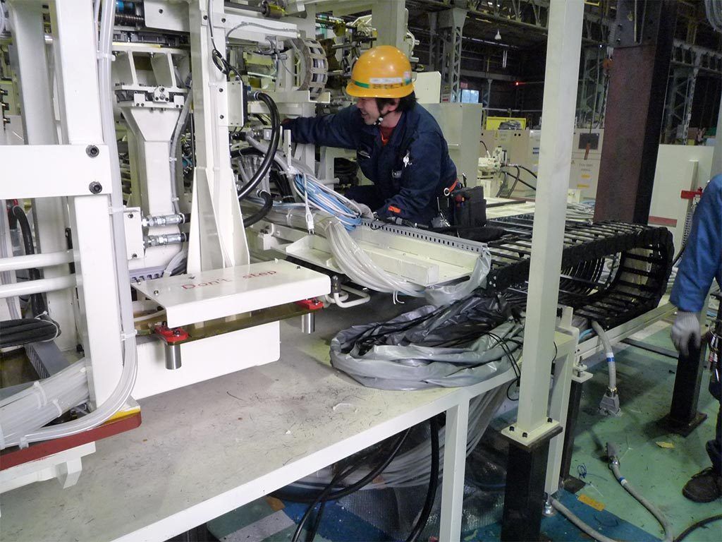 Man in hard hat working on machinery in a factory; black cable carrier visible.