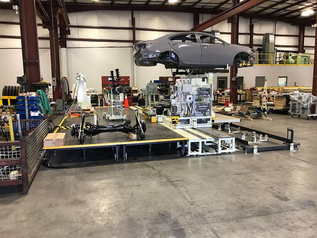 Car body suspended over assembly line in a factory, parts and equipment surrounding.