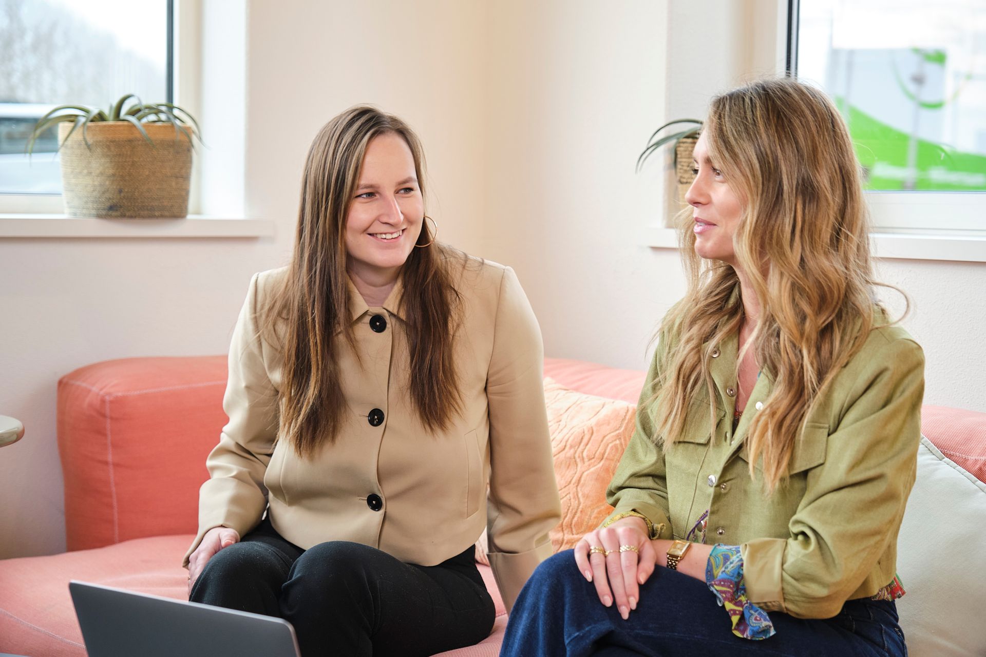Twee vrouwen zitten op een roze bank, lachend en pratend in een lichte kamer.